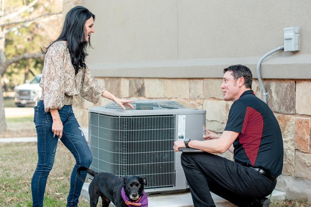 Woman interacting with a Trane HVAC unit alongside a technician, with a dog nearby, highlighting expert service and customer engagement in HVAC maintenance.